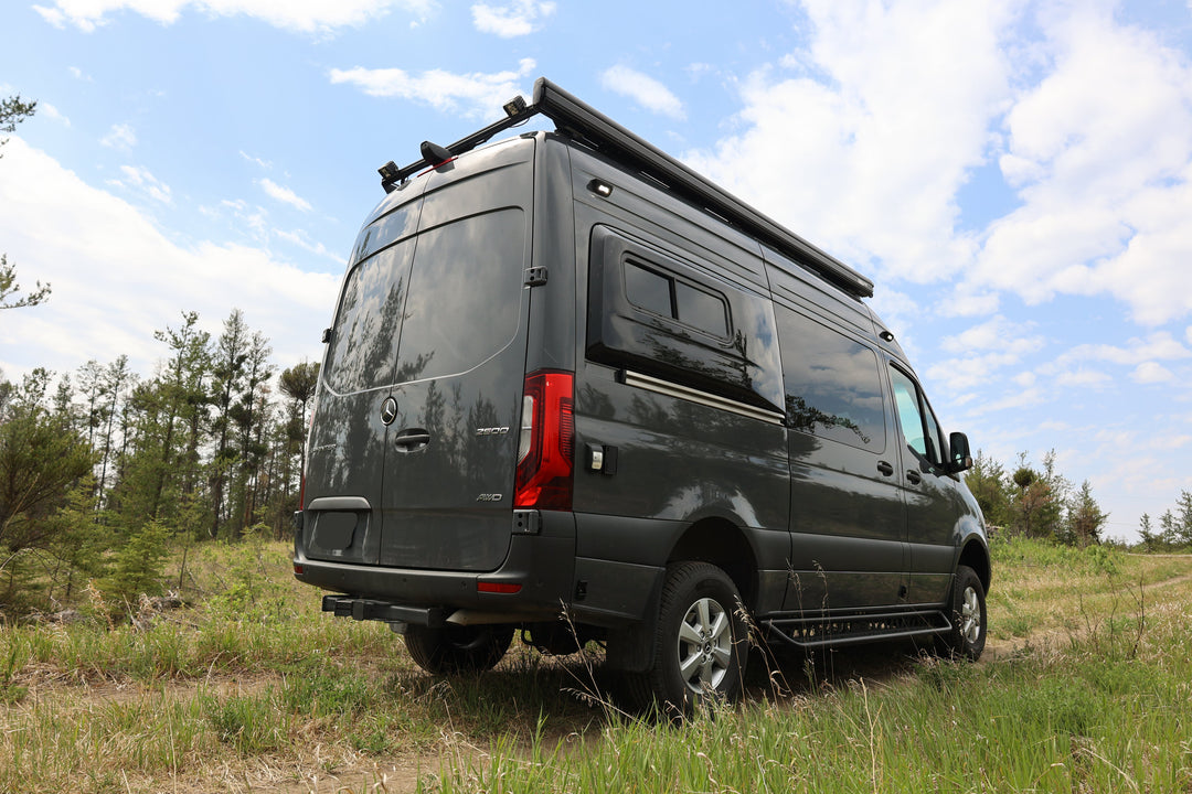 gray sprinter van conversion with a roof rack in a grassy field with trees and blue sky.