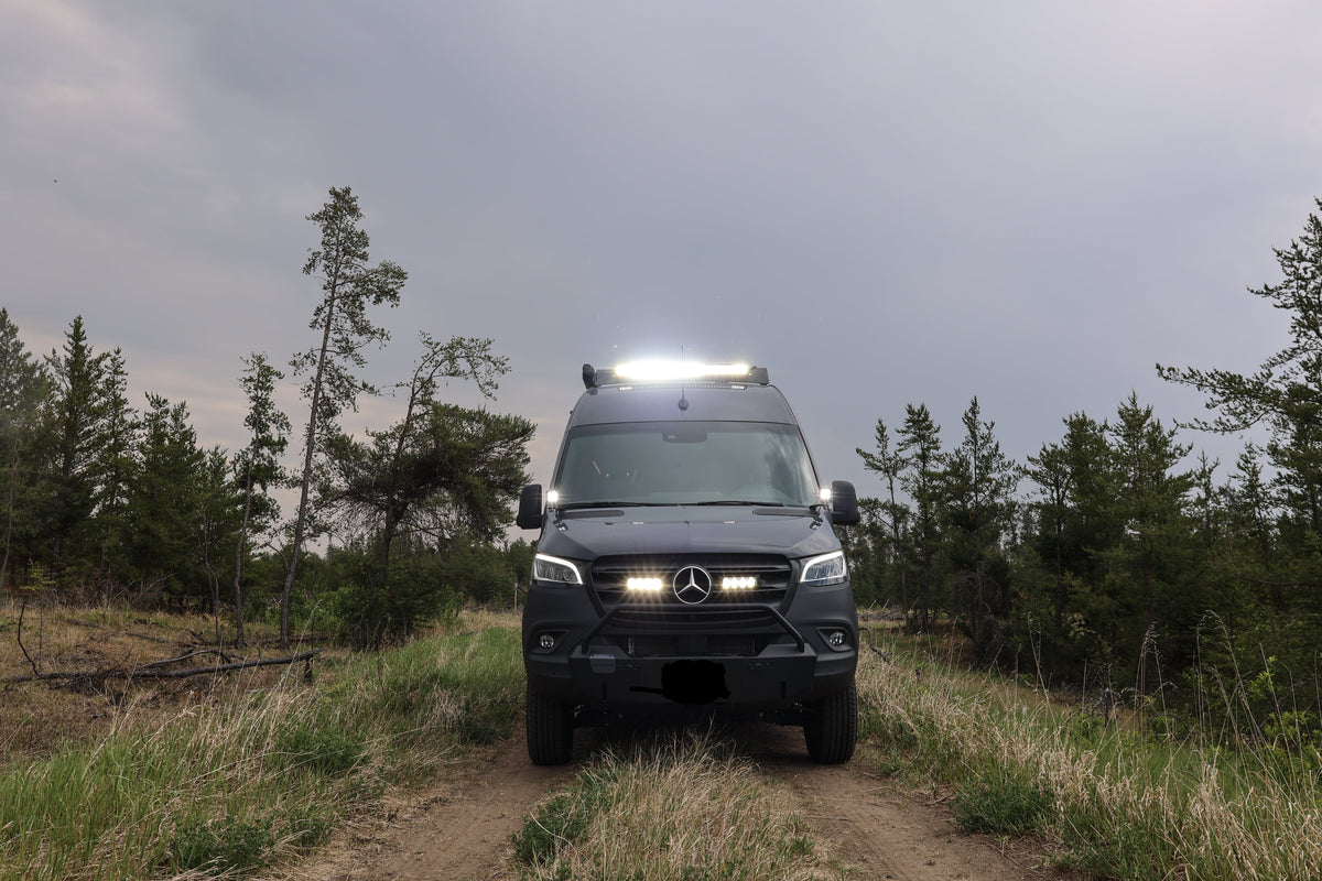Sprinter Van with lights on driving through a forest on a cloudy day